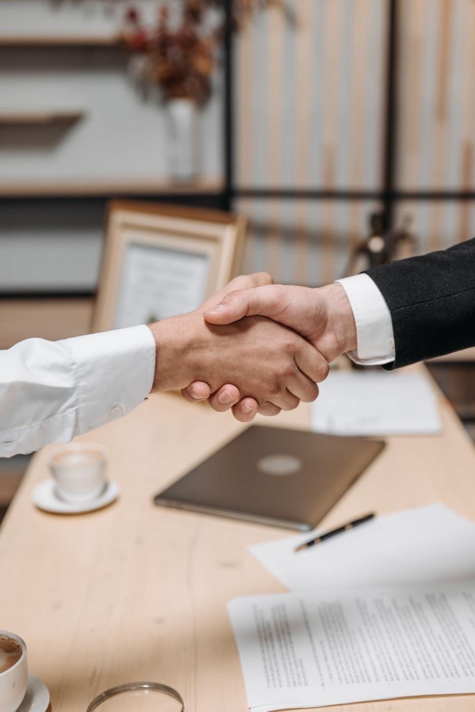 Two people shaking hands over a desk with a laptop and papers